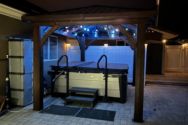 Covered patio with hot tub, wooden pergola, blue accent lighting, and white privacy fence at night